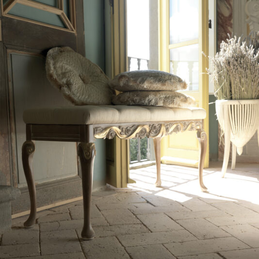 A Designer Rose And Ribbon Italian Bench with intricate carvings stands in front of an open door, bathed in sunlight. Cushions are stacked on the bench, and a large potted plant is beside it. The room has a rustic tile floor and ornate decor, creating a serene and vintage atmosphere.