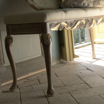 The Designer Rose And Ribbon Italian Bench, adorned with intricately carved details and light upholstery, is positioned near an open door. Sunlight streams through, illuminating the tiled floor and casting soft shadows. The background includes a balcony with a glimpse of greenery outside.