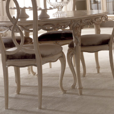 Designer Round Italian Dining Table Set A close-up of the Designer Round Italian Dining Table Set placed on a light-colored carpet. The table features a carved wooden design with ornate legs and a glossy surface. Surrounding the table are cushioned chairs with elaborately carved backs, all in a matching light wood finish.