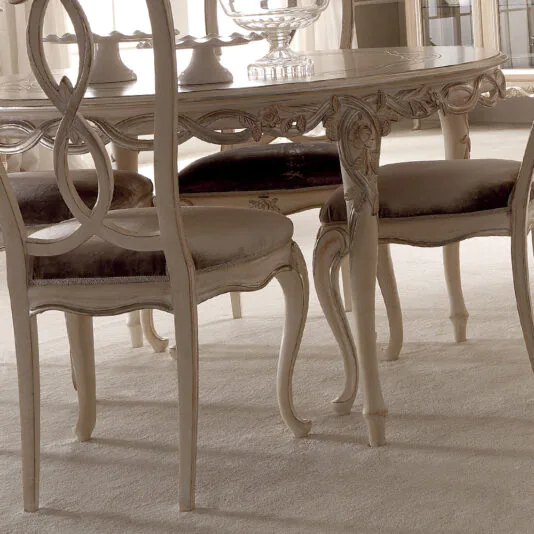 Designer Round Italian Dining Table Set A close-up of the Designer Round Italian Dining Table Set placed on a light-colored carpet. The table features a carved wooden design with ornate legs and a glossy surface. Surrounding the table are cushioned chairs with elaborately carved backs, all in a matching light wood finish.