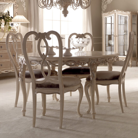 A sophisticated dining room showcasing the Designer Round Italian Dining Table Set accompanied by four ornate, high-back chairs with intricate designs. The room is complemented by a chandelier, a decorative cabinet with glass doors, a side table, and elegant soft lighting. The decor features predominantly white and beige tones.