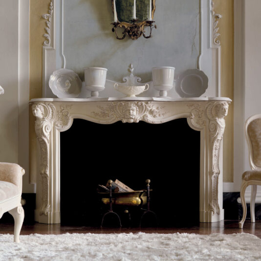 An elegant ivory-colored fireplace featuring intricate carvings stands against a decorative wall, prominently showcasing the Elaborate Italian Fireplace Surround. The mantel is adorned with two white vases and a white urn, while a luxurious beige armchair and plush white rug complete the sophisticated setting.