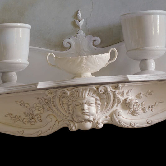 The Elaborate Italian Fireplace Surround, with its ornate white shelf featuring intricate floral carvings and a sculpted face at the center, exudes elegance. To enhance its classical design, a white ceramic urn and bowl are symmetrically displayed on either side of the mantel.