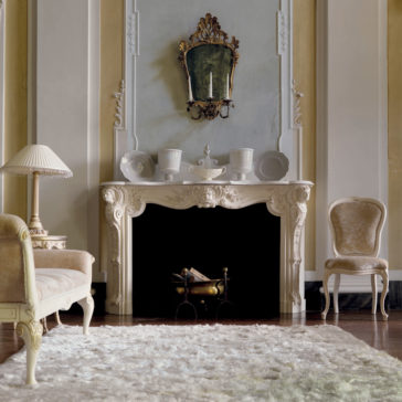 Elaborate Italian Fireplace Surround Experience an elegant living room with a vintage feel, featuring an Elaborate Italian Fireplace Surround with ornate white decorative carvings and an antique mirror above it. White china beautifully adorns the mantel. A cream sofa and two matching chairs provide comfortable seating, while a plush white rug covers the hardwood floor. A classic table lamp adds a touch of soft lighting to complete the ambiance.