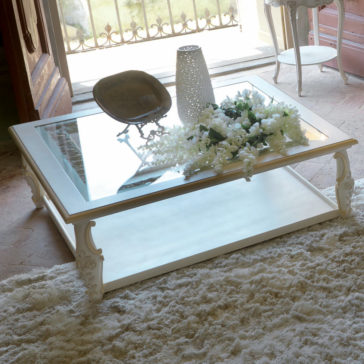 Exclusive Italian Square Coffee Table The Exclusive Italian Square Coffee Table, featuring a glass top and ornate white legs, showcases a decorative arrangement of white flowers in a delicate white vase. Beside the vase is a bronze dish. The table is placed on a plush beige rug, with sunlight streaming through the balcony door in the background.