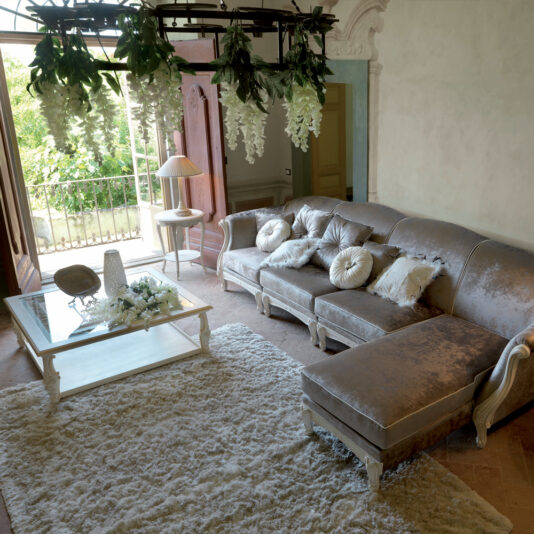 Exclusive Italian Square Coffee Table A luxurious living room features a plush, silver sectional sofa with white throw pillows, a large, fluffy white rug on the floor, and an Exclusive Italian Square Coffee Table adorned with decorative items. A chandelier with hanging greenery is above, and a door opens to a sunny balcony.