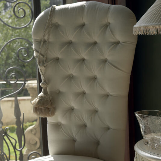 High Button Backed Designer Italian Chair An elegant High Button Backed Designer Italian Chair, featuring a tufted cream upholstery and a decorative tassel draped over the side, is positioned near a sunlit window adorned with an ornate wrought iron railing. A glass lampshade can be seen partially on the right.