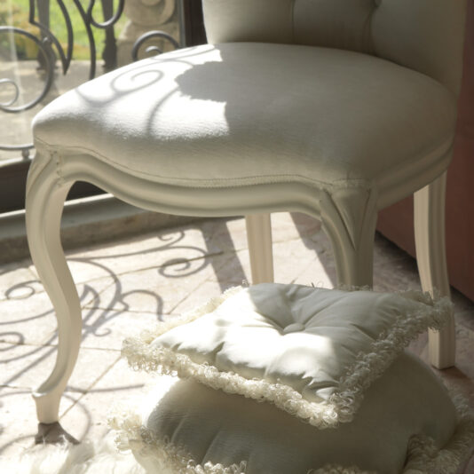 High Button Backed Designer Italian Chair A High Button Backed Designer Italian Chair with elegantly curved legs sits by a sunlit window. Two plush white cushions with decorative fringes are stacked on the floor next to it, basking in the natural light that casts gentle shadows on the surface.