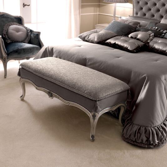 An elegant bedroom with muted tones features a plush, button-tufted headboard on the bed, adorned with matching pillows and a silky bedspread. At the foot of the bed is an ornate Italian Designer Bedroom Bench. A cozy armchair with decorative cushions sits by the window.
