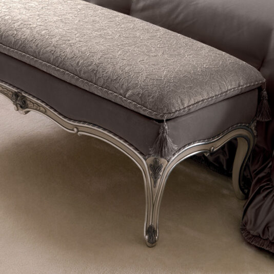 Italian Designer Bedroom Bench A close-up photo of the Italian Designer Bedroom Bench showcasing its beige, intricately patterned cushion. The bench features carved wooden legs with a silver finish and tassel decorations at the edges, set against draped fabric in a matching color.