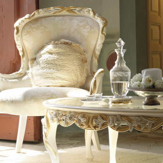 A luxurious setting featuring the Italian Rose And Ribbon Armchair adorned with a textured white and gold cushion. In front of the chair is a matching ornate table displaying a crystal decanter, a white cup on a saucer, flowers, and a small bowl. Sunlight streams in from the left.
