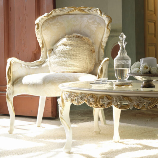 A luxurious setting featuring the Italian Rose And Ribbon Armchair, characterized by its ornate white and gold design, intricate detailing, and plush cushion. In front of the armchair stands a matching elegant coffee table, adorned with a crystal decanter, glasses, and a plate of pastries. The room is softly lit.