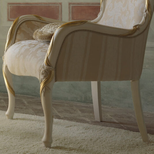 The Italian Rose And Ribbon Armchair, featuring opulent patterned beige fabric, elaborate gold accents, and delicately carved rose designs on its legs, is set atop a light-colored rug in front of a beautifully painted decorative wall.