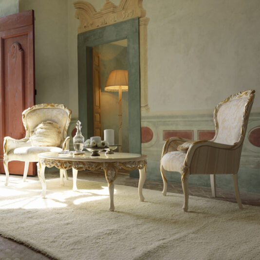 A stylish living room with two elegant Italian Rose and Ribbon Armchairs and a matching coffee table on a plush light-colored rug. A table lamp illuminates the scene from the back corner, with murals and a rustic door adding charm to the sophisticated setting.