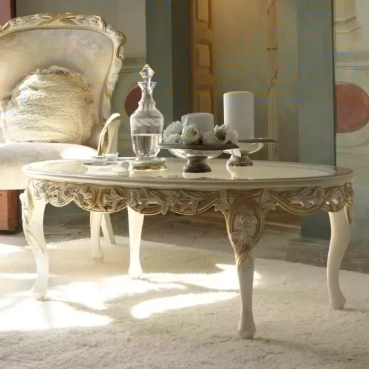 A luxurious living room scene with an ornate Italian Rose And Ribbon Oval Coffee Table in white and gold, featuring intricate floral carvings. On the table, two candles, a decorative tray with small white objects, and a crystal decanter are elegantly arranged. In the background stands an elegant upholstered chair.