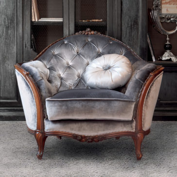 An elegant Louis Button Upholstered Armchair in gray velvet with a tufted backrest and wooden frame sits on a gray carpet. A round, gray tufted throw pillow rests on the seat. Behind the chair is a dark wooden cabinet filled with books and decorative items, complementing the sophisticated scene.