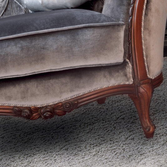 Close-up of an elegant vintage Louis Button Upholstered Armchair with a wooden frame and ornate carvings. The plush velvet upholstery appears worn, adding to its classic charm. The carpet beneath is a soft, neutral tone, complementing the antique furniture.