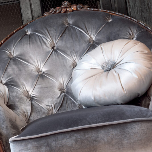 A close-up of the Louis Button Upholstered Armchair reveals its luxurious texture complete with button details and a richly carved wooden frame along the top edge. The armchair is adorned with a round, decorative pillow that features a shimmery, pleated design with a central embellishment.