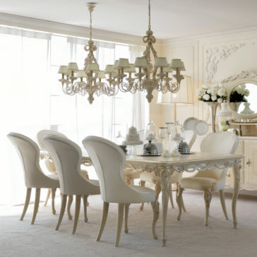 Luxurious Designer Italian Dining Chair A sophisticated dining room with vintage charm showcases an intricate chandelier hanging above a beautifully carved wooden table set for six, complemented by Luxurious Designer Italian Dining Chairs. The space is adorned with large windows draped in sheer curtains, a sideboard displaying a floral arrangement, and soft beige carpeting.