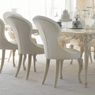 Luxurious Designer Italian Dining Chair A set of Luxurious Designer Italian Dining Chairs, with their elegant off-white hue and curved backs upholstered in textured fabric, are arranged around a matching table in a bright room bathed in soft natural light. The table is beautifully adorned with silver dishware, glassware, and a tiered white cake.