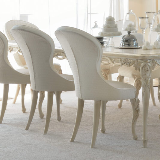 Luxurious Designer Italian Dining Chair A set of Luxurious Designer Italian Dining Chairs, with their elegant off-white hue and curved backs upholstered in textured fabric, are arranged around a matching table in a bright room bathed in soft natural light. The table is beautifully adorned with silver dishware, glassware, and a tiered white cake.