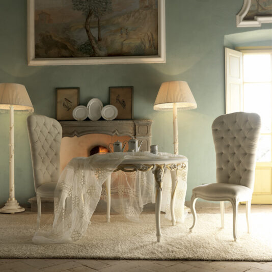 A cozy vintage dining scene features two tufted chairs and a **Luxurious Designer Round Italian Dining Table Set** adorned with a lace tablecloth, ready for tea. A fireplace with decorative plates, two framed artworks, and ornate lamps create a warm ambiance, accentuated by a large painting above the fireplace.