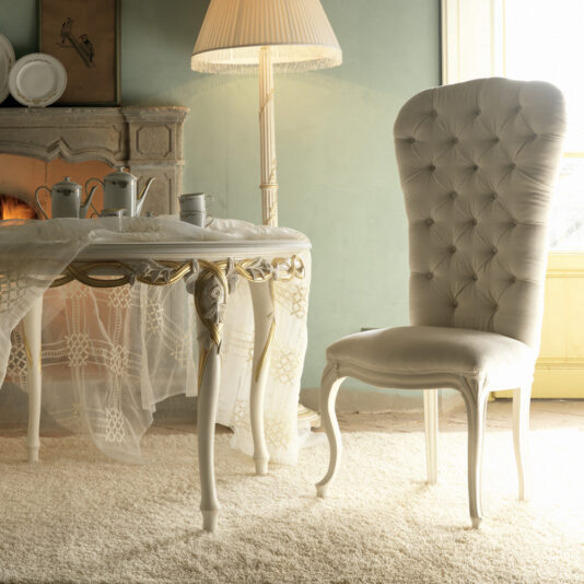A vintage-themed room showcasing an elegant high-backed tufted chair and a Luxurious Designer Round Italian Dining Table Set adorned with a lace tablecloth, tea set, and cups. A lit floor lamp paired with a fireplace adds to the cozy ambiance, while sunlight streaming through the window enhances the warm atmosphere.