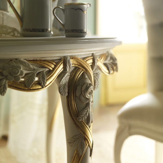 Close-up of the Luxurious Designer Round Italian Dining Table Set featuring intricate floral carvings and gold accents. On this ornate piece, there is a ceramic pot and creamer with a blue rim. The background is softly lit, suggesting a bright room with pastel-colored decor.