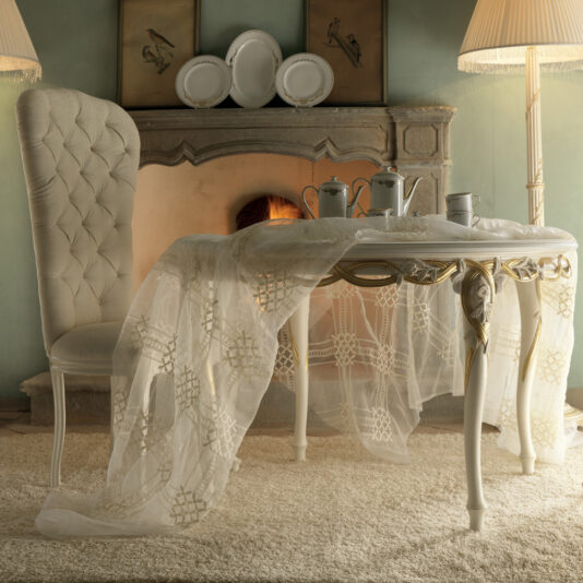 A cozy dining scene showcasing the Luxurious Designer Round Italian Dining Table Set, elegantly draped with a delicate lace tablecloth and adorned with silver tea service items. Beside it stands a tufted chair, while a fireplace with a roaring fire and framed bird artwork above provides the perfect backdrop.