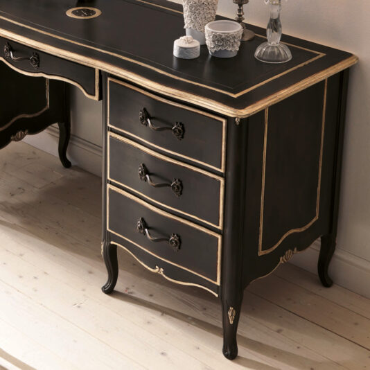 The Luxurious Italian Desk in vintage black with gold trim showcases ornate handles and intricate carvings on its three drawers and legs. Its desk surface elegantly displays decorative items including a lace-covered container, a glass candlestick, and a small, refined bowl.