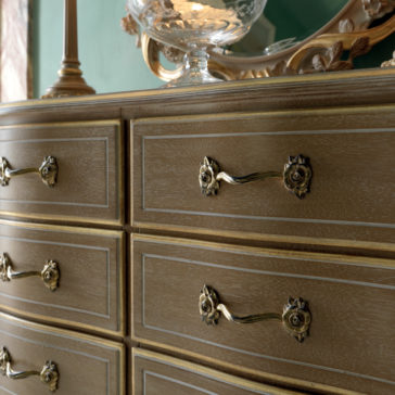 Luxurious Italian Small Chest of Drawers A close-up shot showcases the Luxurious Italian Small Chest of Drawers, featuring six ornate gold-handled drawers. A decorative lamp and a carved mirror are partially visible on top. The dresser boasts a light wood finish with a delicate grain pattern, exuding elegance and craftsmanship.