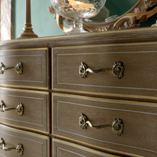 A close-up shot showcases the Luxurious Italian Small Chest of Drawers, featuring six ornate gold-handled drawers. A decorative lamp and a carved mirror are partially visible on top. The dresser boasts a light wood finish with a delicate grain pattern, exuding elegance and craftsmanship.