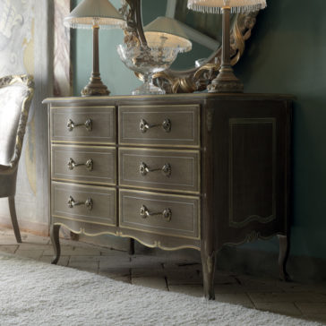 Luxurious Italian Small Chest of Drawers The Luxurious Italian Small Chest of Drawers with ornate handles stands against a green wall. This vintage dresser boasts six drawers and features intricate carvings. Adorned on top are two elegant table lamps with fringe shades and a large, detailed crystal bowl.
