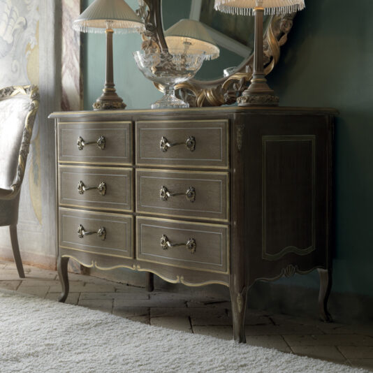 The Luxurious Italian Small Chest of Drawers with ornate handles stands against a green wall. This vintage dresser boasts six drawers and features intricate carvings. Adorned on top are two elegant table lamps with fringe shades and a large, detailed crystal bowl.