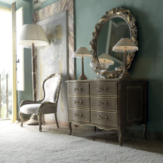 A luxurious room featuring a Luxurious Italian Small Chest of Drawers with six compartments, topped with two elegant table lamps and a decorative bowl. A large round mirror with a detailed frame hangs above. A cushioned armchair and a tall floor lamp stand nearby, with a balcony door open.