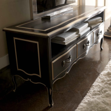 Luxurious Louis Italian TV Unit The Luxurious Louis Italian TV Unit is a vintage black stand with decorative off-white trim, featuring three open shelves that hold stacks of books and three closed drawers adorned with ornate metal handles. Resting on curved legs, this elegant piece is placed on a dark polished floor next to a white rug.