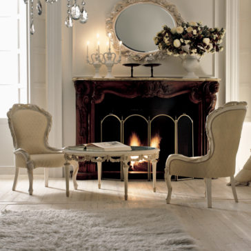 A cozy, elegant living room features an ornate fireplace adorned with candles, a large mirror, and a floral arrangement on the mantel. Two Luxury Italian Rose And Ribbon Armchairs are set around a small table with an open book. A plush rug covers the light wooden floor.