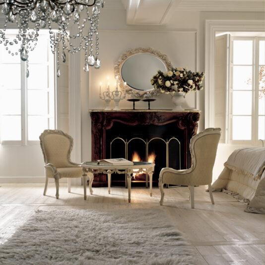 Luxury Italian Rose And Ribbon Armchair Refined living room with vintage-style decor, featuring two Luxury Italian Rose and Ribbon Armchairs placed around a small table in front of a lit fireplace. Above the fireplace is a round mirror and an elegant floral arrangement. A crystal chandelier hangs from the ceiling, while natural light streams through large windows. A plush white rug covers the wooden floor.