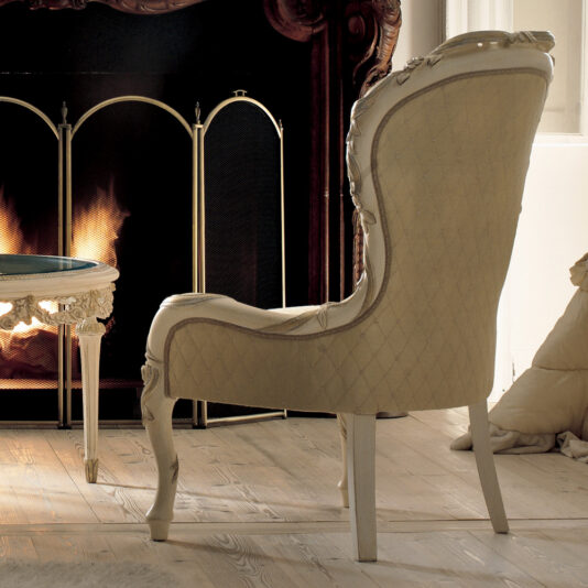 Luxury Italian Rose And Ribbon Armchair A Luxury Italian Rose and Ribbon Armchair with plush, quilted upholstery sits facing a lit fireplace. The chair features carved detailing and is positioned on a wooden floor. A small side table is partially visible to the left, while warm light from the fire illuminates the cozy room.
