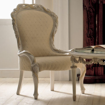 An elegant, vintage cream-colored chair with intricate carvings on the backrest and legs stands next to a similarly styled table in a well-lit room with light wooden floors. This Luxury Italian Rose And Ribbon Armchair complements the open book resting on the table perfectly.
