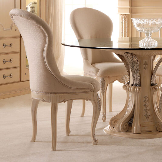 An Opulent Italian Dining Chair with a curved backrest is positioned beside a round glass table. The table boasts an ornate, carved pedestal base and is set within a classic, lavishly decorated room. Another identical chair can be seen partially in the background.