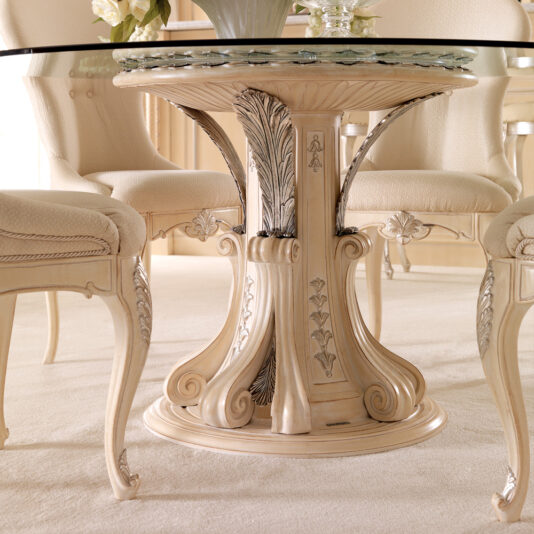 A close-up of the Opulent Italian Round Glass Dining Table Set showcases its exquisitely carved pedestal base adorned with floral and leaf motifs. Surrounding the table are three cream-colored chairs featuring elaborate leg and backrest designs, all elegantly placed on a beige carpet.