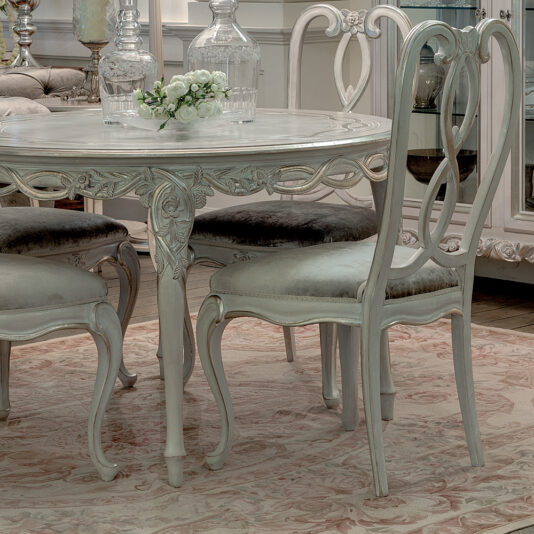 Ornate Designer Round Italian Dining Table Set The Ornate Designer Round Italian Dining Table Set, featuring a glass-top table and elegantly crafted chairs, is placed on a patterned rug. The table is beautifully decorated with a small flower arrangement and glass decanters, embodying a vintage style characterized by intricate detailing and a soft, sophisticated color palette.