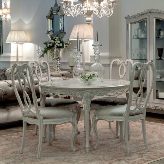 Elegant dining room with vintage decor featuring an Ornate Designer Round Italian Dining Table Set and four ornate chairs. Two crystal decanters and a small flower arrangement sit on the table. A chandelier hangs above, and a mirrored cabinet and wall mirror are in the background.