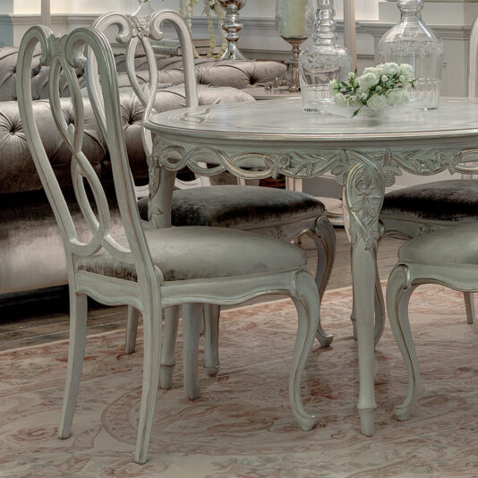 Ornate Designer Round Italian Dining Table Set A vintage-style dining room showcases the Ornate Designer Round Italian Dining Table Set, adorned with a glass decanter and a small bouquet. The setting includes elegant decor, white chairs, a cushioned sofa in the background, and a patterned rug covering the floor.