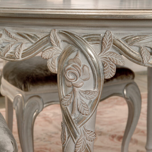 Ornate Designer Round Italian Dining Table Set Close-up of an ornately carved leg from the Ornate Designer Round Italian Dining Table Set, showcasing intricate rose and leaf designs with a gently weathered silver finish. The matching chair is partially visible in the background, upholstered in lush silvery-gray fabric, enhancing the elegant interior.