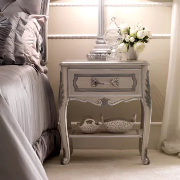Ornate Italian Bedside Table An Ornate Italian Bedside Table graces the bedroom with its beautifully intricate design, exuding a classic, vintage style. This elegant piece holds a glass jar and a vase filled with white flowers, while its lower shelf is neatly organized with two baskets. The surrounding decor complements this arrangement with light, pastel colors.