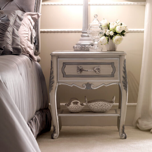 An Ornate Italian Bedside Table graces the bedroom with its beautifully intricate design, exuding a classic, vintage style. This elegant piece holds a glass jar and a vase filled with white flowers, while its lower shelf is neatly organized with two baskets. The surrounding decor complements this arrangement with light, pastel colors.