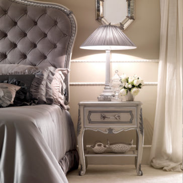 Ornate Italian Bedside Table A luxurious bedroom showcases a tufted headboard, the Ornate Italian Bedside Table adorned with intricate carvings, a table lamp featuring a pleated shade, a vase of white flowers, and a decorative bowl on the lower shelf. The decor is dominated by soft, neutral tones, creating an ambiance of serene elegance.