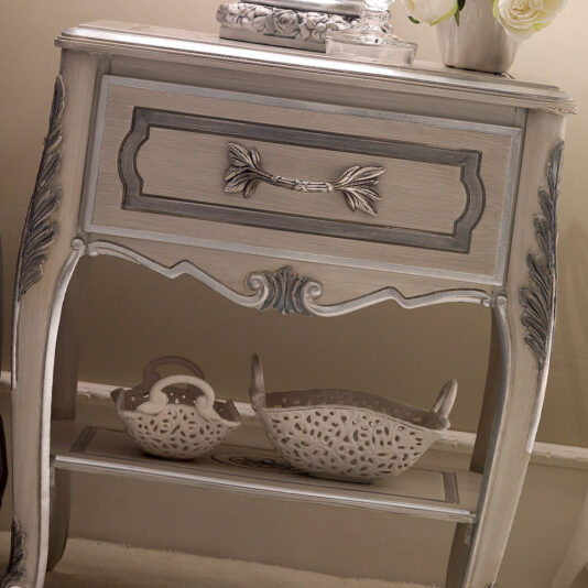 The Ornate Italian Bedside Table is a vintage-style piece featuring intricate carvings and a weathered gray finish. It includes a single central drawer adorned with elaborate details. The lower shelf showcases two decorative white baskets with lace-like patterns, while the tabletop is beautifully accented with a gray vase filled with flowers.